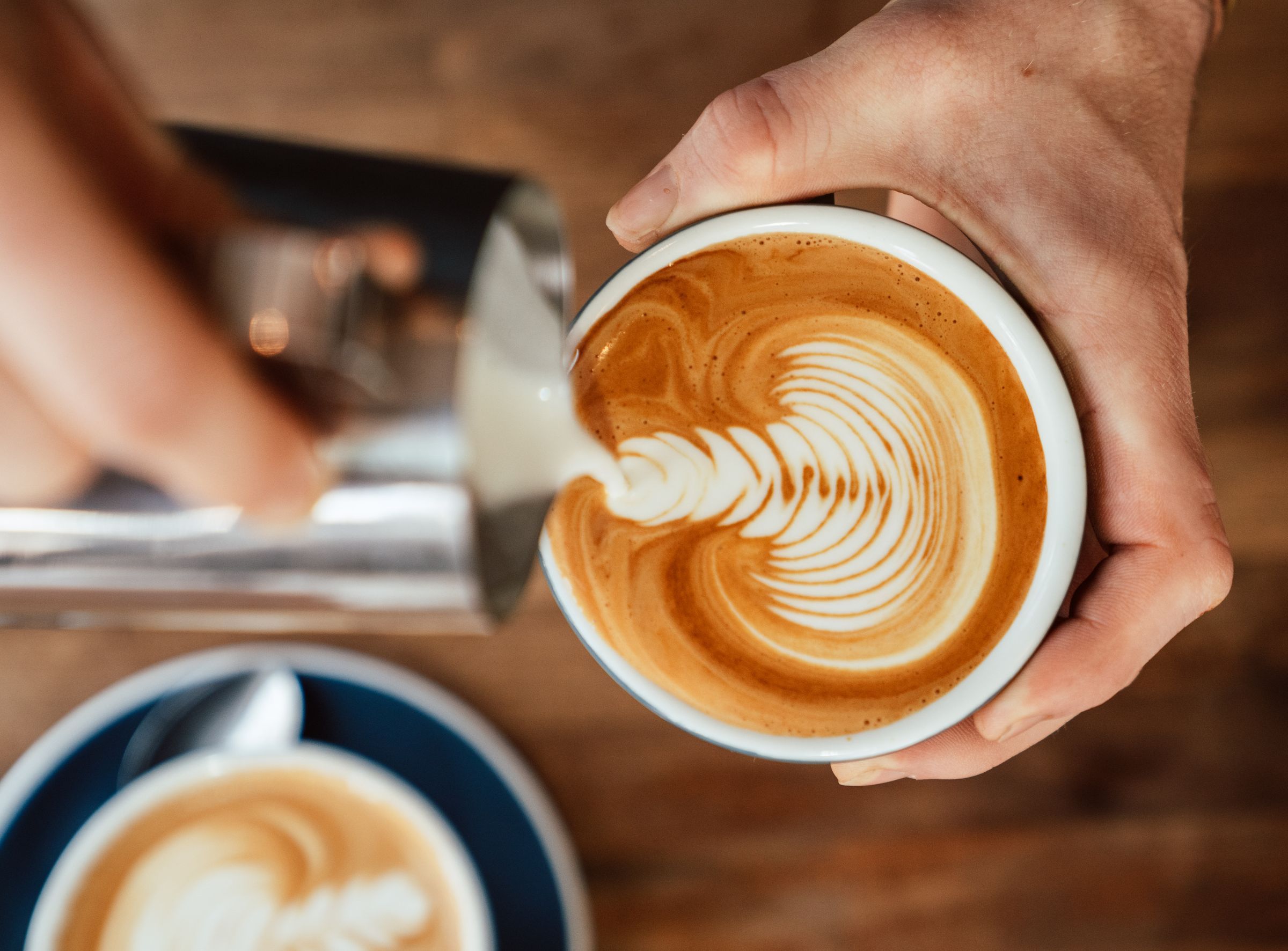 Latte Art von oben: Milchschaum wird in eine Tasse gegossen, gehalten von zwei Händen